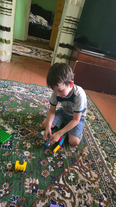 A young boy is playing with building blocks on a rug in a room. He is sitting on his knees and focused on the blocks in his hands. The rug is green with a floral pattern. In the background, there is a doorway leading to another room with a bed, and a television set on a wooden cabinet. The floor is made of wooden planks. The scene appears to be in a residential setting.