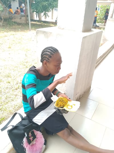 A person with braided hair is sitting on a tiled surface in Urualla, Nigeria. The individual is wearing a horizontally striped teal, black, and white long-sleeved shirt, with black shorts, and is holding a white foam plate of food, which appears to be noodles with fried plantain. They are reaching out with their right hand towards the food. A black bag is resting next to them on the tiled floor. The person is sitting next to a concrete column. In the background, there are trees, grass, and a building. Other people can be seen in the background as well.