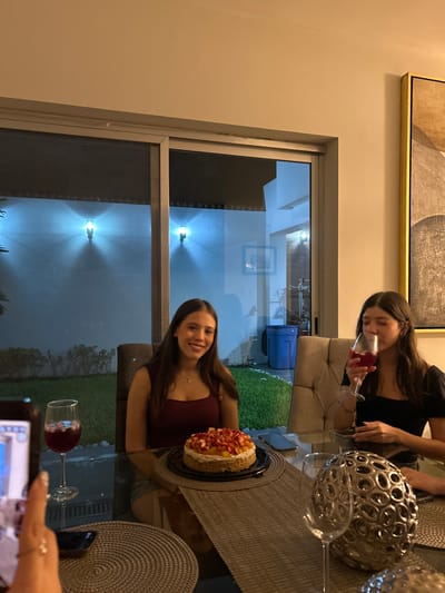 Two women are seated at a glass table, likely indoors near a window. One woman is drinking from a wine glass, while the other is smiling and seated near a cake decorated with strawberries. An elaborate decorative metal ball sits on the table, along with wine glasses. A cellphone is visible in the foreground, possibly taking a photo. Through the window, exterior lighting and glimpses of vegetation are visible. The location is speculated to be in Tampico, Mexico.