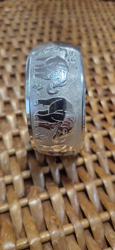 A close-up, slightly angled shot shows a silver bracelet with an embossed design of two elephants. The elephants are facing each other, with their trunks curled. The bracelet is resting on a woven surface, possibly a basket or mat, made of light brown material. The background is out of focus, emphasizing the detail of the bracelet. There is no visible indication of the location in Póvoa de Varzim, Portugal.