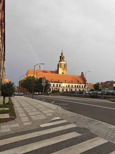 The image depicts a street scene in Stargard, Poland. In the background, a large building with a prominent tower and red-tiled roofs dominates the view. A faint rainbow is visible in the overcast sky above. In the foreground, a sidewalk with a patterned stone surface leads to a crosswalk. The street is paved with asphalt, and parked cars are visible along the side. A lamppost stands on the sidewalk. The overall impression is a quiet urban environment, possibly after a rain shower, given the presence of the rainbow.