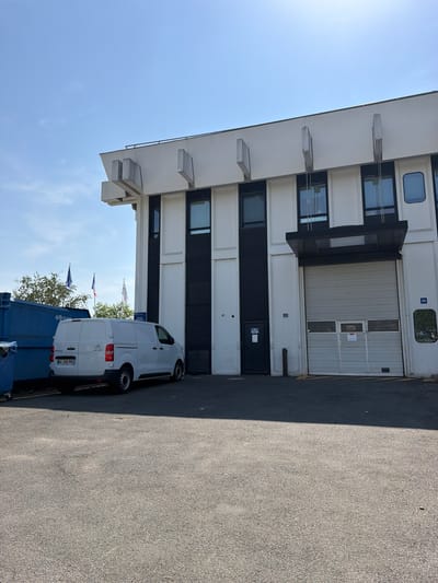 This media file captures an outdoor scene in Rungis, France, likely during daytime given the bright sunlight and shadows. The primary subject is a modern, white commercial van parked in front of a multi-story building with a large, ribbed garage door. The building has a clean, minimalist design with several windows, some rectangular and some with rounded corners. One of the windows has the number "55" next to it, and another smaller door with the same number is visible below the garage door.
The van, a white panel van, is positioned to the right of the frame, facing away from the building. Its rear license plate is partially visible, showing "GL 310 PE". Partially obscured behind the van is a dark blue dumpster.
In the background, beyond the van and dumpster, are trees and several flags flying on poles. The sky is a clear, bright blue, suggesting fair weather. The ground appears to be asphalt. The overall scene is functional, likely depicting a commercial or industrial area, with no people visible. The time of day is suggested by the strong shadow cast by the building onto the asphalt, indicating a sunny period.