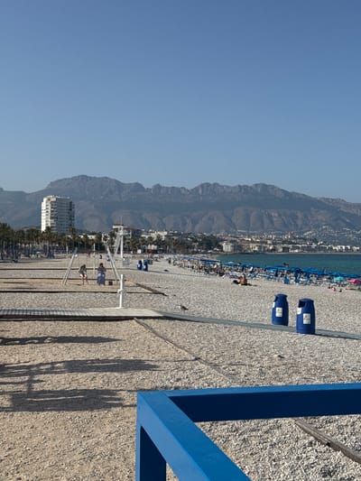 This image depicts a sun-drenched beach scene in Pla Parcial Platja, Spain. The foreground is dominated by a gravelly beach area with a wooden walkway and some sand. Two blue trash receptacles are visible. In the middle ground, a playground with swings is set up on the sand, with two individuals on the swings. Beyond the playground, rows of blue beach umbrellas and lounge chairs line a section of the beach, with people scattered among them. Palm trees are visible on the left side of the image. The background features a cityscape with buildings, including a tall white high-rise, and a prominent mountain range under a clear blue sky. The Mediterranean Sea is visible on the right side of the image, with calm water. The overall impression is one of a peaceful, recreational beach environment.