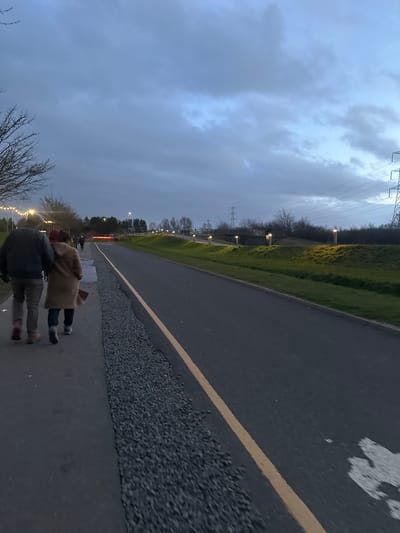 The image depicts a multi-use path and adjacent road at dusk within Helix Park, Falkirk, United Kingdom. Two individuals walk away from the camera on a dark grey paved path on the left. The person on the left wears a dark jacket, light trousers, and a dark cap. The person on the right wears a light brown long coat and a red hat, carrying a dark bag. A strip of grey gravel separates the paved path from a darker grey asphalt road on the right, which features a single yellow painted line.
To the left of the path, bare tree branches are visible, with strings of small, illuminated white lights draped overhead. Further down the path, indistinct figures are visible in the distance. Along the right side of the road, a green grassy embankment slopes upwards, illuminated by a series of cylindrical, upward-glowing bollard lights spaced at regular intervals. Beyond the embankment, dark trees and bushes are present. A faint reddish-orange glow is visible on the road in the mid-distance, and a dark structure, possibly an underpass or bridge, is seen beneath the embankment, also with illumination. In the far background, multiple tall metal electricity pylons and power lines stand against an overcast, blue-grey sky.