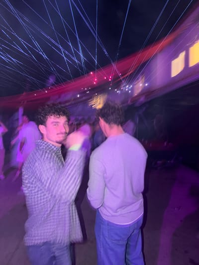 Two people are captured in a blurred, rotated image. One person wears a gray sweater and blue jeans. The other wears a plaid shirt and jeans, looking toward the camera with curly hair. The background includes a structure with many thin blue lines stretching across a dark sky, illuminated with pink and white lights. The overall ambiance is purple and chaotic.
