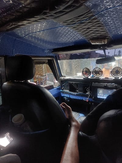 The image depicts the interior of a vehicle, likely a taxi or private car, situated in an urban environment in Mwandege, Tanzania. The perspective is from a passenger seat, looking forward towards the dashboard and windshield. The vehicle's ceiling is upholstered with a patterned blue fabric and black quilted sections. A bare, tanned leg extends across the front passenger seat. A white cup with a brown interior is visible in a side pocket of the front left seat. The dashboard, primarily blue, holds three small, circular electric fans with decorative black grills, one exhibiting a red glow. A digital screen, possibly displaying a music interface, is visible on the right side of the dashboard, accompanied by several wires. Through the front windshield, a blurred exterior street scene is visible, featuring buildings and indistinct figures, consistent with a bustling urban area. A mobile phone with a brightly illuminated screen is partially visible in the lower left corner, held by the individual whose leg is shown.