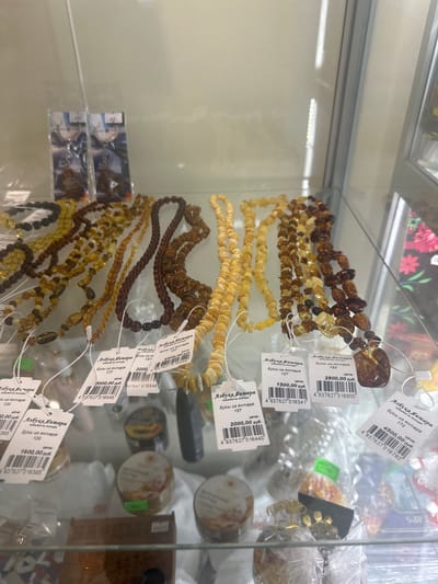 This media file captures a close-up view of several necklaces made of amber beads, displayed in a glass cabinet. The necklaces vary in color, ranging from light yellow to deep brown, and are arranged in cascading layers. Each necklace has a small tag attached, displaying Cyrillic text and a barcode. The text on the tags appears to indicate the product type ("Бусы из янтаря" - amber beads), a price (e.g., "2800,00 руб." - 2800.00 rubles, "1500,00 руб." - 1500.00 rubles), and potentially a product code or model number. The overall setting is a retail environment, likely a jewelry store or market stall, with other products visible in the background, some of which are also in packaging. The lighting is bright, suggesting an indoor setting during daylight hours. The focus is clearly on the amber jewelry, highlighting its natural variations in color and form. The scene is static, with no people visible, emphasizing the merchandise. The location context provided is Lyambir', Russia.