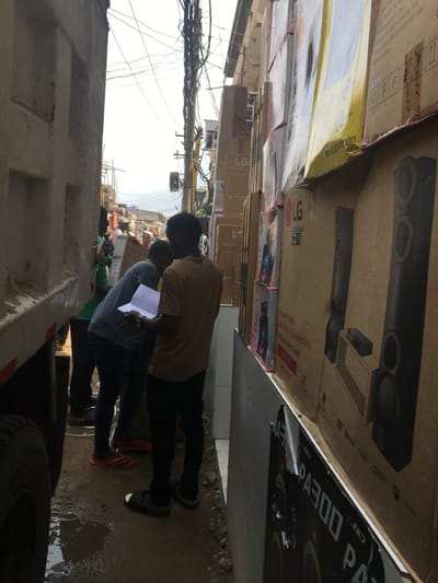 The image displays a bustling, narrow commercial passageway in Ojo, Nigeria, under an overcast sky. The ground consists of wet, uneven earth with visible puddles and debris. On the left, the light-colored side and a large tire of a utility or delivery truck occupy the foreground. The right side of the passage is a wall covered entirely by stacked cardboard boxes of various sizes, predominantly featuring LG brand electronics, including televisions and large floor-standing speakers.
In the mid-ground, three men are visible. One man, wearing a tan t-shirt and dark trousers with dark sandals, stands facing away from the camera, holding an open notebook or clipboard and looking down at its contents. Slightly to his left and forward, a second man in a blue/grey long-sleeved shirt, dark trousers, and red flip-flops is bent over, seemingly inspecting an item near the ground, while also appearing to observe the notebook. A third individual, partially obscured by the truck on the left, wears a green shirt and is handling a large, flat box.
The background reveals a densely packed urban environment with various structures, utility poles, and numerous entangled overhead electrical wires, indicating a busy market or commercial district. Multiple indistinct figures are also visible further down the passage.