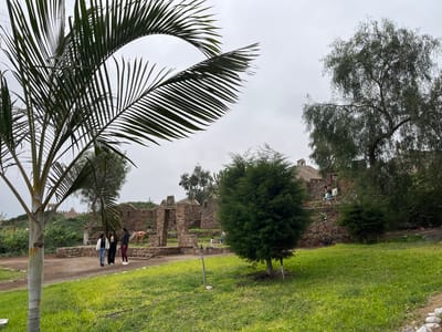 The image shows a park in Quilmaná, Peru. Three women walk along a path near stone ruins. A palm tree is in the foreground, partially obscuring the view. Grass covers the ground, and other trees dot the landscape. Overcast skies suggest a cloudy day. People are visible on the ruins, as well as animal sculptures.