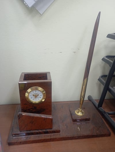 The image shows a desk set on a wooden surface. The desk set consists of a pen holder with a clock on its front and a pen with a gold-colored tip. The pen holder is made of a dark wood-like material with a marble pattern. The clock has a white face with Roman numerals and a double-headed eagle emblem, typical of the Russian coat of arms. The pen holder also has a slanted piece of the same dark material in front of it. The pen itself is long and slender with a wooden shaft and a gold-colored metal tip and base. In the background, a portion of a white wall is visible, and to the right, a black metal shelf unit is partially in frame. The setting appears to be an indoor desk or workspace. There is no information in the image to indicate the location is the city of Ararat, Armenia.