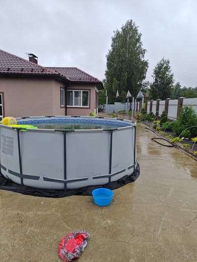 The image depicts an outdoor scene in Vorsma, Russia, featuring a large, above-ground swimming pool filled with water. A yellow and blue inflatable ball and a green inflatable ring are visible floating in the pool. The pool is situated on a concrete patio, which appears wet, suggesting recent rain. A small blue plastic basin filled with water is placed on the patio near the pool, and a crumpled red and black patterned fabric item lies on the ground nearby.
In the background, a tan-colored house with a dark brown tiled roof and a chimney is visible. Adjacent to the house, a section of a white and brick fence runs along the property line, with several small evergreen bushes and flowering plants planted along its edge. A black garden hose is coiled on the wet patio in front of the garden beds. The sky is overcast and grey. The scene suggests a residential setting, likely a backyard, during damp weather.