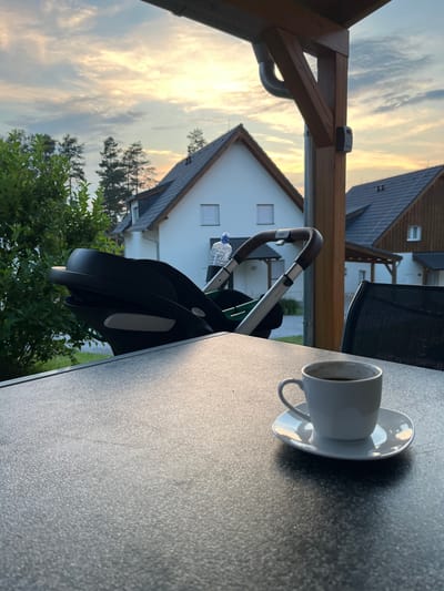 A horizontal, close-up shot captures a white coffee cup and saucer on a dark, speckled table. The cup is half-filled with a dark liquid, likely coffee. In the background, a baby stroller is partially visible, with its brown leather handles and silver frame. A clear plastic bottle of water is tucked behind the stroller. Beyond the stroller, houses with dark roofs and white walls are visible, along with lush green trees and bushes. The sky is a blend of soft yellow, orange, and blue, indicating either sunrise or sunset. The overall atmosphere suggests a moment of quiet relaxation on a patio or balcony. There is no visible text in the image.