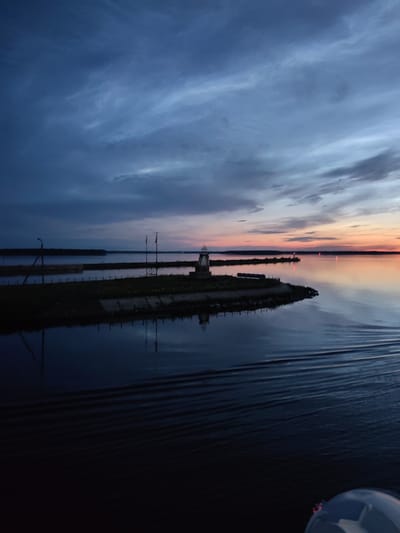 The image depicts a body of water, possibly a river or a lake, at dusk. The sky is overcast with dramatic clouds in shades of blue and grey, with a hint of orange and pink near the horizon, indicating the setting sun. In the foreground, the water is dark and reflects the sky and the surrounding landscape. There are visible ripples on the water's surface, suggesting movement, perhaps from a boat.

On the left side of the image, a dark silhouette of land with some vegetation is visible, with a tall, thin structure, possibly a lamppost, on the edge. In the mid-ground, a small, man-made peninsula juts out into the water. It is topped with a small structure that resembles a lighthouse or a memorial, and there are what appear to be flags or poles. The peninsula is lined with a low wall or embankment, and there are some scattered lights visible on the water in the distance.

The overall impression is one of a serene, yet dramatic, natural landscape during twilight. Given the information that the location is Sheksna, Russia, this could be a view of the Sheksna River or one of the nearby bodies of water. The scene captures the quiet beauty of the environment at the end of the day.