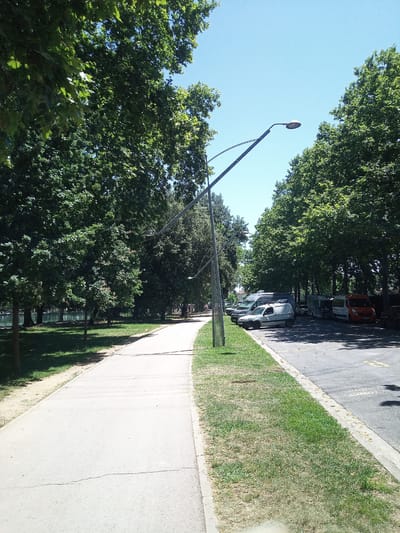 This media file depicts a sunny day in a park in Banyoles, Spain. The scene features a paved walkway curving to the right, bordered by a lush green grassy area on its left and a parking lot on its right. Mature trees with dense green foliage line both sides of the walkway and parking lot, casting dappled shadows. A modern streetlamp with a curved design is prominently featured, extending over the walkway and parking area. Several vehicles are parked in the lot, including vans, a camper, and an orange van with text on its side that is not legible. In the background, a body of water, likely a lake or river, is visible to the left, with more trees on its far bank. A few distant figures can be seen walking along the path or in the park area. The clear blue sky suggests fair weather and daytime. There are no discernible emotions conveyed in the image, and the focus is on the natural and urban elements of the park.