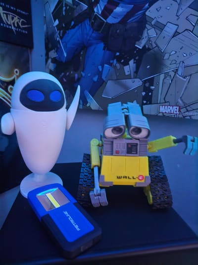 The image shows a display of objects on a dark surface, illuminated by a dominant blue light. Central to the scene are two robot toys: a yellow and grey WALL-E with tracks and the text "WALL·E" on its side, and a white, oval-shaped EVE with blue-detailed "eyes." A blue and black flashlight, branded "PETROLINE" and emitting a bright yellow-white light, stands to the right of EVE.

In the background, a comic book featuring a Captain America-like character amidst debris is visible, with "MARVEL" printed near the top. Below EVE, part of a book or media case shows text fragments including "IS MY STORY" and "RESURRECTION." The dim, blue ambient light creates a cool, display-like atmosphere. The stationary objects suggest a personal collection or decorative arrangement in an indoor setting in Alto Hospicio, Chile. No specific activity or event is taking place; the items are static.