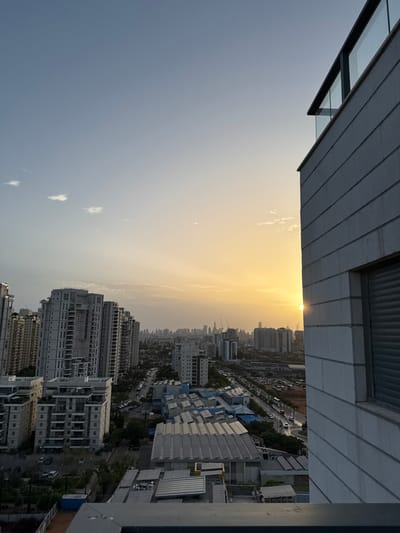 The image shows a cityscape of Ganei Tikva, Israel, as seen from a rooftop. The sun is setting in the distance, casting a warm glow on the city. The image is taken from a building with a white brick facade and a large window on the right side. The rooftop is visible in the foreground, with a railing and a glimpse of a tennis court. Several buildings are visible in the background, with trees scattered throughout the city. The scene is quiet and peaceful, with no people visible.