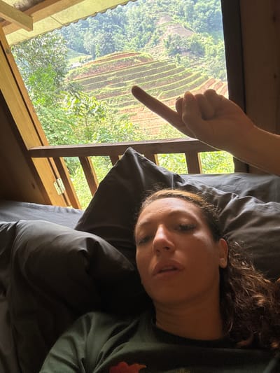 The image shows a person lying on a dark gray pillow and bedding in an indoor space, likely a room in a building. A hand with a pointing finger is positioned above the room's window, which offers a view of the rice terraces landscape in the Hoàng Su Phì Commune, Vietnam. The window frame appears to be made of wood. Outside, the terraced fields are visible, along with lush green trees.