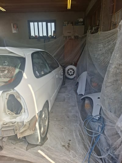 The image shows an indoor garage scene in Rakitovo, Bulgaria. A white vehicle, appearing to be a car, is partially covered with translucent plastic sheeting. Components of the vehicle are scattered around the area on the floor, including a tire and an aftermarket rim. A blue hose is also present next to these components. In the background, the garage wall is visible, with a window on the left and storage shelves at the top-left. The garage appears to be in the process of bodywork and/or painting.