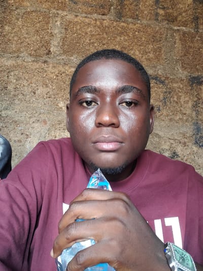 A portrait-style photograph, taken in Gboko, Nigeria, features a young man with dark skin, short dark hair, and a neatly trimmed beard, looking directly at the camera. He wears a maroon long-sleeved t-shirt, with partially visible white text on its front displaying the characters "1" and "T". His expression is calm and direct.
He holds a clear plastic bottle of water in his left hand, positioned near his face. The bottle has a blue label, but the specific text on it is obscured by his grip. On his left wrist, he sports a distinctive silver-toned watch with a large, ornate green face and visible blue accents, possibly gems.
The background presents a rough, textured surface, likely a concrete or brick wall, characterized by earthy tones and dark speckles, suggesting an outdoor or semi-outdoor environment. The lighting appears natural, indicating it is daytime. The image captures a moment of the man holding water, perhaps for hydration, against a rugged backdrop.