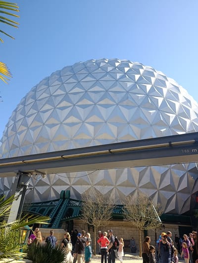 The image depicts a large, geodesic sphere-shaped structure made of numerous triangular panels, likely constructed of metal or a similarly reflective material. The structure appears to be part of an amusement park ride or attraction, with a monorail track running horizontally across the middle of the building. The text "3.60 m" is visible on the monorail.
At the base of the structure, a crowd of people is present, suggesting that the structure is a popular attraction. The people are of varying ages and are dressed in casual clothing. Some appear to be standing or walking, while others are standing in groups.
In the foreground, there is some greenery, including palm-like plants. The sky is clear and blue.
Given the style of architecture and rides, it is likely that the location is Europa-Park in Rust, Germany.
