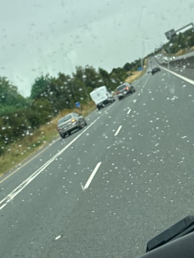 The image captures a view from inside a vehicle on a highway, likely during rainy weather, as indicated by the water droplets on the windshield and the wet appearance of the road. Multiple vehicles are visible on the multi-lane road, including a silver sedan, a white van, and a dark-colored car further ahead. The surrounding environment consists of lush green trees and vegetation lining the sides of the highway. A road sign is partially visible in the distance. The scene depicts typical highway traffic conditions, possibly in or near Father Campion Place, Ireland, given the context provided.