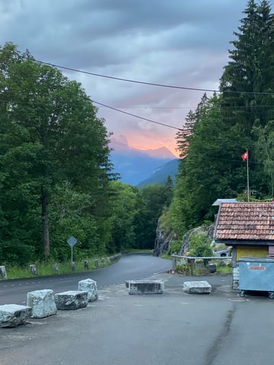 This image depicts a mountainous, forested landscape in the vicinity of Innertkirchen, Switzerland, during either dawn or dusk. The sky above is largely overcast with grey clouds, but a horizontal band of orange and pink light from the setting or rising sun illuminates distant, silhouetted mountain peaks in the upper-middle background. The scene is dominated by dense green foliage, with tall coniferous and deciduous trees lining both sides of a dark asphalt road that curves gently to the left and ascends into the background.

In the foreground, a paved area features five rough-hewn, light grey stone blocks arranged in a line, acting as barriers. The road itself appears wet, reflecting the dim light, and has a dashed white line marking its right edge. Along the left side of the road, a line of short, grey stone posts serves as a barrier, and a diamond-shaped road sign is partially visible.

On the right side of the image, adjacent to the road, stands a small building with an orange-brown tiled roof. A red Swiss flag with a white cross on a pole is positioned to the right of this building. Next to the building, a large, grey metal dumpster with German text is visible on wheels. A wooden fence runs behind the stone blocks and in front of the building. Utility lines, dark in color, stretch horizontally across the upper third of the frame. No people or active vehicles are present in the scene.