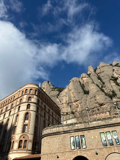 The image captures a panoramic view in Collbató, Spain, featuring mountainous terrain and architectural structures under a blue sky with scattered white clouds. The upper right and background are dominated by the imposing, light grey and tan conglomerate rock formations of the Montserrat mountains, exhibiting characteristic rounded peaks and steep faces, with sparse green vegetation clinging to crevices. In the mid-ground left, a multi-story building with a rounded corner is visible, constructed with alternating cream-colored plaster sections and reddish-brown brickwork. It features multiple arched windows on each of its five visible floors, and a white cylindrical object is atop its roof. In the lower right foreground, a stone-block structure with a curved facade presents several large, vertically oriented windows containing graphic banners that partially spell "MUS" and "EU" from "MUSEU". Three dark-colored streetlights are mounted along the upper edge of this foreground structure. Further up the mountainside, behind the foreground structure, smaller, light-colored stone buildings with arched windows are discernible on a terrace. No individuals or active subjects are present. Direct sunlight illuminates the scene, casting shadows on the left side of the prominent multi-story building.