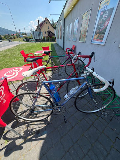 The image shows two bicycles parked outside a building in Międzybrodzie Bialskie, Poland. The building appears to be a store or restaurant, with posters displayed on its exterior wall. Red tables and chairs are set up on a paved area in front of the building, suggesting an outdoor seating area. One bicycle is blue and the other is red. Both bicycles are leaning on a green bike rack. The weather is sunny and clear, with a bright blue sky and some clouds. In the background, there are trees, houses, and hills, indicating a semi-rural or suburban setting. The bicycles are the primary subjects, and they are parked and unattended. There are no people visible in the image.