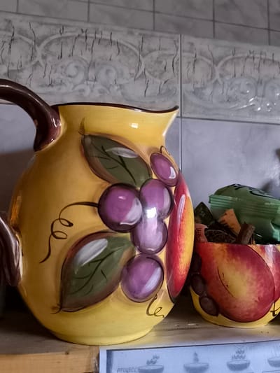 In Žiaukos, Lithuania, on a shelf in what appears to be a kitchen or dining area, two decorative ceramic containers are visible against a tiled backsplash. The larger item on the left is a yellow jug adorned with a painted motif of purple grapes and green leaves. To its right, a smaller, bowl-shaped container is filled with various items, including what appear to be tea bags and possibly cinnamon sticks. This container is decorated to resemble a red and yellow fruit, likely an apple. The overall scene suggests a domestic setting, likely a kitchen, with an emphasis on decorative kitchenware. The background shows a light-colored, possibly stone or ceramic tile backsplash with a repeating pattern.