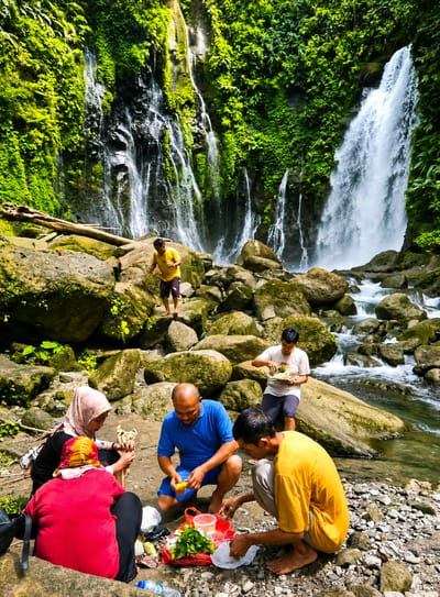In Gunung Ambat, Indonesia, a group of five people is gathered near a cascading waterfall. The waterfall, with its numerous streams, descends from a lush green cliff face covered in vibrant foliage. A river flows in the foreground, its water rushing over rocks.

Four individuals are seated or crouched on the rocky ground, engaged in preparing and eating food. One person, wearing a blue t-shirt and shorts, is peeling a fruit. Another, in a yellow t-shirt, is also preparing food. A person wearing a pink hijab and a black top is holding some food items. A fourth individual, in a red shirt and yellow headscarf, is seated in front of them, also with food. A fifth person, standing near the edge of the rocks in the background, is also holding food. The group appears to be having a picnic. Scattered around them are various food items, including vegetables, fruits, plastic bags, and a bottle of water. The overall atmosphere is relaxed and convivial, set against the backdrop of the natural beauty of the waterfall and its surroundings.