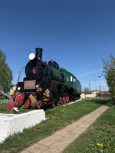 The image shows a stationary steam locomotive displayed on a white stone base in Balezino, Russia. The locomotive is black and green with red accents on the wheels. Wreaths and floral arrangements surround its base. A small plaque is affixed to the front of the locomotive. The sky is clear and blue. A sidewalk and green grassy area are adjacent to the display. Other buildings are visible in the background.