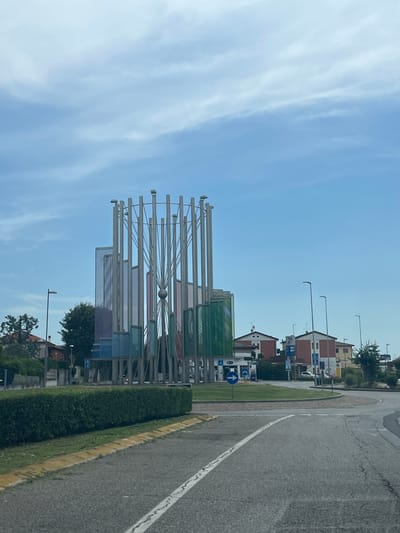 The image shows a modern art sculpture in the center of a roundabout in Castiglione delle Stiviere, Italy. The sculpture is made of metal poles and colored glass panels, with a spherical element in the center. In the background, there are residential buildings, a gas station, and streetlights. The sky is cloudy. The roundabout is paved with asphalt and has lane markings. A neatly trimmed hedge lines the edge of the roundabout.