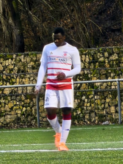 A soccer player in a white jersey with red stripes stands on a green field. He wears white shorts and orange cleats. Behind him is a stone wall and some trees. The field has white lines marking it. The player appears to be looking down, perhaps at the ball or the ground. The jersey has a sponsor logo visible.