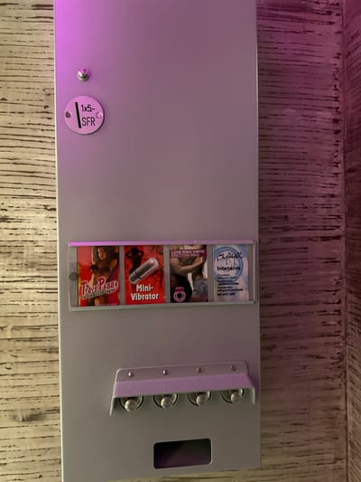 A close-up, high-angle shot captures a gray, industrial-style vending machine or dispenser, likely located indoors in Winterthur, Switzerland. The machine is mounted against a textured wooden wall, which appears to be composed of vertical planks. A distinct purple or pink light emanates from the left, casting a soft glow on the machine's surface and the upper portion of the wall.
The primary subjects are various adult novelty products displayed within clear, vertical slots on the right side of the machine. From top to bottom, these products are:
* A package of "Oyaya Intensive" condoms, with text noting "3 kondome transparent feucht," "3 préservatifs transparents lubrifiés," "3 preservativi trasparenti lubrificante," and "ISO 4074:2002."
* A product labeled "LOVE RING VIBRO ERECTION RING."
* A "Mini-Vibrator," depicted with a silver, bullet-shaped device.
* A "Travel Pussy Die künstliche Vagina."
To the right of the product display, a silver metal bracket holds four visible metallic knobs or buttons, arranged vertically, which likely correspond to the selection of each product. Further right, a dark, rectangular opening serves as a retrieval slot for dispensed items. On the lower-left portion of the machine, a small, circular silver tag is visible, engraved with "1x5- SFR" and a vertical line, indicating a price point of 5 Swiss Francs. A small silver screw or bolt is also present next to this tag. The machine's overall appearance suggests it is a discreet dispenser for adult items.