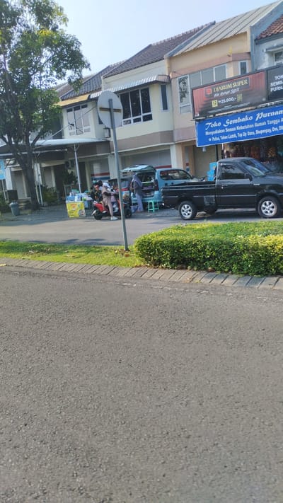The image depicts a street scene in Sidodadi, Indonesia, on a clear day. In the foreground, a paved road with a distinct curb runs diagonally from the bottom left to the top right of the frame. Adjacent to the road is a neatly trimmed green hedge. Behind the hedge, a sidewalk area is visible, with a traffic signpost standing vertically near the center.
Further back, a row of buildings lines the street. The buildings have light-colored facades with a cream and beige color scheme, featuring tiled roofs and awnings over some windows. Several storefronts are visible, with some shutters closed. A large advertisement poster for "DIJARUM SUPER" is prominently displayed on one building. Another banner below it announces "Toko Sembako Purnam" (Purnam's Grocery Store) and lists various services including "Menyediakan Semua Kebutuhan Rumah Tangga Anda" (Providing all your household needs) and offers for "Pulsa, Token Listrik, Top Up Dana, Shopeepay, Ovo, Dll" (Airtime, Electricity Tokens, Fund Top Ups, Shopeepay, Ovo, etc.).
On the sidewalk in front of the stores, there is activity. A red scooter is parked, with a person standing next to it. A light blue van is parked behind the scooter, with its rear doors open, revealing shelves stocked with various items. A person is standing next to the open van. In front of the van and scooter, a yellow food cart with a sign is visible, suggesting a food vendor. A few other individuals are present in the vicinity of the scooter and the food cart. A black pickup truck is parked to the right of the blue van. A large green tree with dense foliage is situated to the left of the buildings, casting some shade. The sky is clear and bright blue, indicating sunny weather.