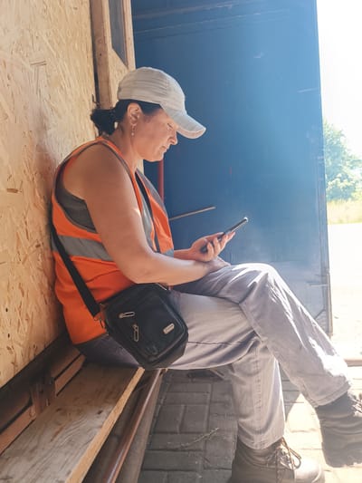 A woman wearing a high-visibility orange vest and a light-colored baseball cap is seated on a wooden bench. She is looking down at her smartphone, which she holds in her right hand. A black crossbody bag is slung over her left shoulder. The woman is wearing light-colored pants and dark boots. She is sitting against a wall constructed of oriented strand board (OSB) and next to a dark blue doorway, which opens to an outdoor area with trees and sunlight. The ground is paved with interlocking bricks. The scene appears to be an outdoor break area, possibly at a construction site or industrial facility. The location is the city of Vuhlehirsk, Ukraine.