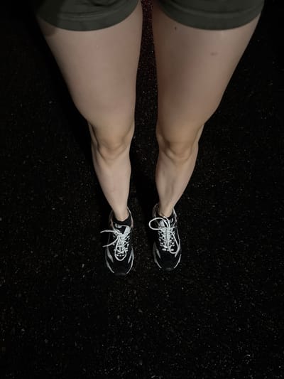 The image shows a low-angle shot of a person's legs and feet, wearing athletic shorts and black and white sneakers. The person is standing on a dark, asphalt surface that appears to be wet or reflective. The lighting is dim, suggesting it might be nighttime or an indoor setting with low light. The background is uniformly dark and out of focus, emphasizing the legs and feet as the primary subjects. There are no discernible landmarks or environmental details that would indicate a specific location in Denver, United States, or any other city. The composition is clean and focused on the lower body.