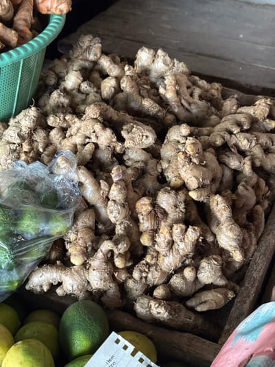 In the city of Karangsambung, Indonesia, an overhead shot reveals a pile of ginger roots displayed at a market stall. The ginger, with its rough, textured skin, is predominantly light brown and tan. Some of the roots are larger and knobby, while others are thinner and more elongated. To the left, a portion of a green plastic basket is visible, containing more of the ginger, along with some orange-hued turmeric roots. A clear plastic bag, partially filled with green limes or small gourds, lies on top of some of the ginger. In the lower portion of the image, several more green limes and a corner of a paper receipt are visible. The background consists of a dark, wooden surface.