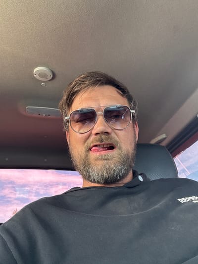 A man is pictured in a selfie, taken from a low angle inside what appears to be a vehicle. The man has a beard and is wearing sunglasses with metal frames. His mouth is open, with his tongue slightly visible. He is wearing a black sweatshirt with white text on the left shoulder that is partially obscured but appears to read "REPR". The background shows the interior of a car, including a grey ceiling with a small circular speaker and a light fixture. Outside the car window, a blurry sky with pink and purple hues is visible, suggesting it may be dawn or dusk. The overall mood is informal and candid.