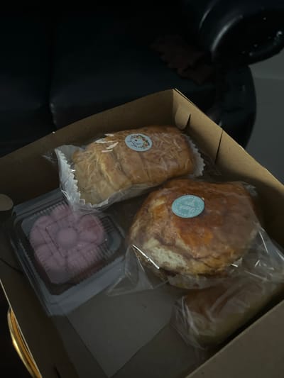 A cardboard box contains four baked goods. Two appear to be loaves of bread, one with a sprinkle of shredded cheese on top and a floral sticker that reads "Thank you". Another loaf has a similar sticker with floral elements. The third item is a square plastic container with pink, layered pastries. The fourth item is partially visible, also a baked good in plastic wrapping. The box is resting on a dark, possibly leather surface, and the background is out of focus, suggesting an indoor setting. The lighting is dim. There is no indication of the city of Jambearum, Indonesia, in the image.