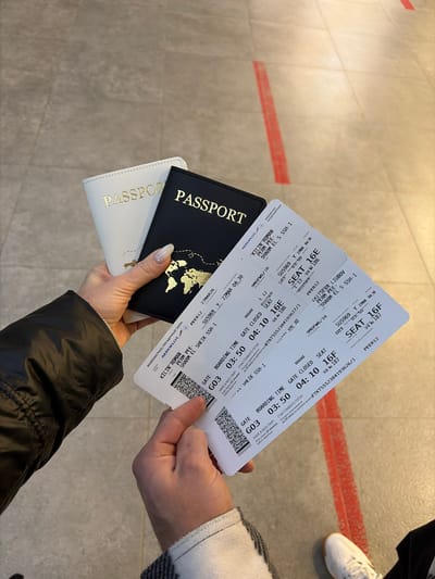 The image displays a close-up, downward-angled view of two individuals' hands holding travel documents within an airport terminal. The left hand, featuring manicured nails with light polish and partially covered by a black puffer jacket sleeve, securely holds two passport covers: one white and one black, both embossed with gold "PASSPORT" lettering and a gold world map graphic. This hand also holds the upper sections of two white paper boarding passes. The right hand, dressed in a grey and white checkered sleeve, grasps the lower portions of the same boarding passes.

The boarding passes, issued by Aeroflot, detail flights for "KILIN ROMAN" (seat 16E) and "ZAITSEVA LIUBOV" (seat 16F) on flight SU5969, scheduled for March 27, 2026. The flight originates from Perm, Russia, and is destined for Sharm El Sheikh, Egypt. Boarding time is 03:50, with the gate (G03) closing at 04:10 for a 04:30 departure. Each pass includes a QR code, flight specifics, and Russian text, notably "СЧАСТЛИВОГО ПУТИ!" (Have a nice trip!).

The floor beneath the hands is composed of light grey tiles with visible grout lines. Two parallel red lines are marked diagonally on the floor, suggesting a designated path or queue. A person's right foot, shod in a white sneaker with dark stripes, is partially visible in the bottom right corner of the frame. The scene captures the moment of preparing for an international flight departing from Perm, Russia, within a modern airport setting. The exact location in the city of Boldino, Russia, cannot be confirmed from the image; the flight originates from Perm.