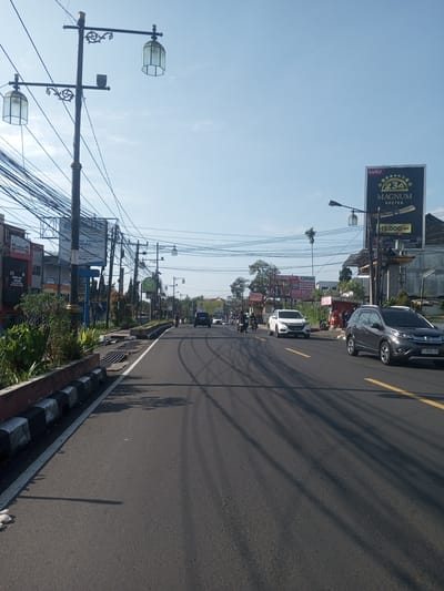 The image depicts a street scene in Nhot Chang, Laos. The street is paved and has tire tracks on it. A grey Honda car is on the right lane. Further down the road, a white car is visible. There are several billboards on both sides of the street, one advertising "MAGNUM KRETEK". Overhead power lines are strung across the street. Streetlights with two ornate lamps are present. Sidewalks are visible on both sides of the street, with plants and buildings bordering the sidewalks. There are motorcycles and other vehicles in the distance. The sky is clear and blue.