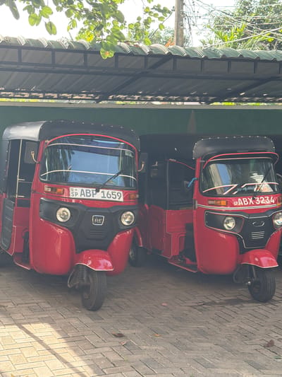 Two red tuk-tuks are parked under a covered structure. The tuk-tuk on the left has license plate "SP ABP 1659". The tuk-tuk on the right has license plate "SP ABX 3234". The ground is paved with interlocking bricks, and some leaves are scattered on the ground. Overhanging branches with green leaves are visible above the covered structure, suggesting an outdoor setting, possibly in a tropical environment like Rumassala, Sri Lanka. The lighting indicates it is daytime. There are no people visible in the image.