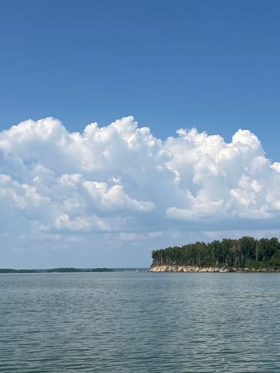 The image depicts a wide expanse of calm, blue water, likely a river or lake, under a bright, clear blue sky. A prominent line of large, fluffy white cumulus clouds stretches across the upper portion of the sky, casting subtle shadows on the water. In the distance, a narrow strip of land is visible, densely covered with a variety of green trees. This landmass features a steep, sandy embankment along its edge. The scene appears tranquil and natural, possibly a waterfront view in or near Bystrovka, Russia, given the context of the prompt. There are no people or specific actions captured in the image, focusing solely on the landscape and atmospheric elements.