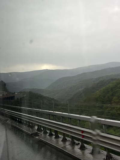 The image depicts a scenic mountain landscape in Gorno Kamartsi, Bulgaria, viewed from a highway overpass. The sky is overcast, suggesting it is daytime with cloudy or rainy weather, as evidenced by water droplets on the visible guardrail. The mountains, covered in lush green trees, recede into the distance, shrouded in a misty haze. The guardrail, made of metal and concrete, runs along the right side of the frame, indicating the perspective of someone traveling on a road. Two small, triangular red markers are visible on the guardrail, potentially for visibility or marking. The overall mood is serene and somewhat somber due to the weather and the vastness of the landscape. No people are visible in the image.