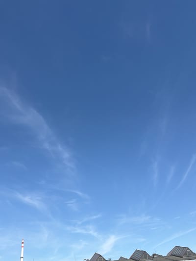 This media file shows a clear blue sky with sparse, wispy clouds. The image is oriented vertically, with the top of the frame pointing towards the sky and the bottom showing a partial view of buildings. On the right side of the frame, the upper edge of several multi-story buildings with distinctive diamond-shaped skylights is visible. A tall, striped industrial chimney with red and white bands is also partially visible in the lower right corner, extending upwards. There are a few small, dark specks in the sky, which appear to be birds in flight. The overall scene suggests an outdoor view from a city, likely during the daytime due to the bright sky and visible structures. The weather appears to be clear and pleasant. No text is visible in the image. The location context is Olomouc, Czechia.