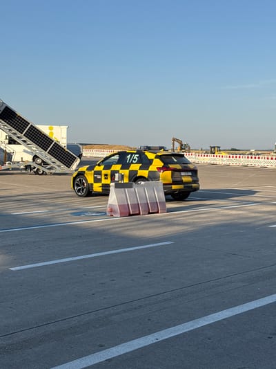An Audi SUV, marked with a black and yellow checkered pattern and the number "1/5" on its rear, is parked on an asphalt surface marked with white lines. The vehicle has the "Memmingen Airport" logo on its side. In the foreground, a series of translucent plastic barriers filled with a reddish liquid are positioned. Behind the car, a mobile vehicle ramp with solar panels is attached to a white trailer. In the background, construction equipment, including an excavator and a bulldozer, are visible along with a white and red striped barrier fence. The scene is set under a clear blue sky.