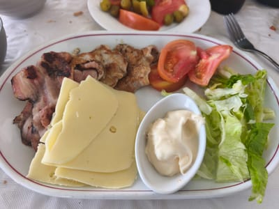 This image captures a meal served on a white tablecloth in Sueca, Spain. The foreground features a white oval plate with a red rim, predominantly displaying a selection of food items. On the left, several strips of grilled bacon with visible char marks are presented alongside multiple slices of grilled meat, possibly pork or chicken. Adjacent to the meats are several overlapping slices of pale yellow cheese. A small, white, boat-shaped bowl contains a serving of creamy, off-white sauce, consistent with mayonnaise or aioli. To the right of the sauce, a mound of shredded green lettuce is visible, followed by several slices of red tomato.
In the background, slightly out of focus, a round white plate contains a side dish of green olives, sliced red tomatoes, and yellow pickled peppers. A silver dinner fork with four tines rests on the white tablecloth to the right of the main plate. A portion of a grey cup or bowl is partially visible in the upper left corner. The tablecloth exhibits a textured, possibly quilted, pattern. No individuals or active interactions are depicted in the scene. The lighting suggests an indoor or shaded outdoor dining environment.