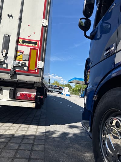 The image shows a blue semi-truck, partially filling the frame, with a white trailer attached. The truck appears to be at a gas station in Niederwinkling, Germany. The sky is visible, with some clouds, and the gas station canopy has a blue border. The rear of the trailer has red and white lights and various stickers, including a yellow "Attention Angles Morts" (Blind Spots) sticker. The scene includes other trucks parked in the distance and a paved surface where the truck is parked. Shadows are visible on the ground.