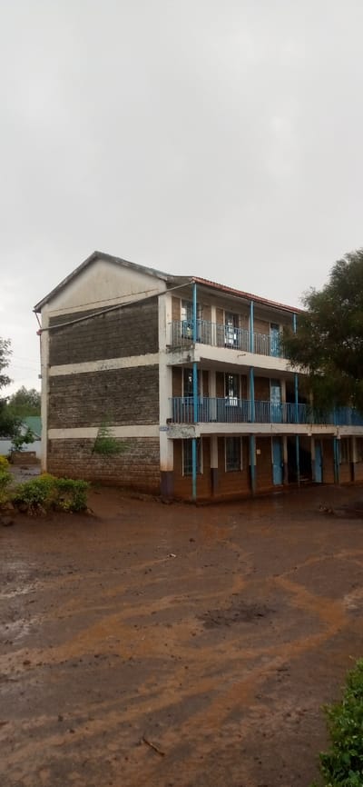 The image displays a three-story building, likely a school or dormitory, set against an overcast sky. The ground surrounding the building is unpaved, appearing very muddy, wet, and reddish-brown, suggesting recent rainfall. Visible tracks and ruts are imprinted in the soft soil.

The building itself features exposed concrete block construction on its left side, with alternating bands of grey blocks and lighter, possibly plastered, horizontal strips. The front facade, facing the viewer, is lighter in color, possibly painted, with a darker brown or reddish base on the ground floor. The building has two upper floors with balconies that span the length of the visible front. These balconies are supported by pillars and feature blue metal railings. Numerous windows and doors, many also painted blue, are visible on each level. The roof on the left side is gabled, with a visible white concrete end, while the roof over the balconies appears to be a flat or slightly sloped overhang. A drainpipe runs down the left side of the building. The overall condition of the building shows signs of wear, with some weathering and dirt accumulation on the walls and balconies.

In the foreground, to the left, there are small green bushes. A larger tree with dense, dark green foliage partially obscures the right side of the building. The sky is a uniform light grey, indicating heavy cloud cover or an overcast day, which aligns with the muddy ground conditions. No identifiable text is visible in the image. The scene appears to be set during daytime.
